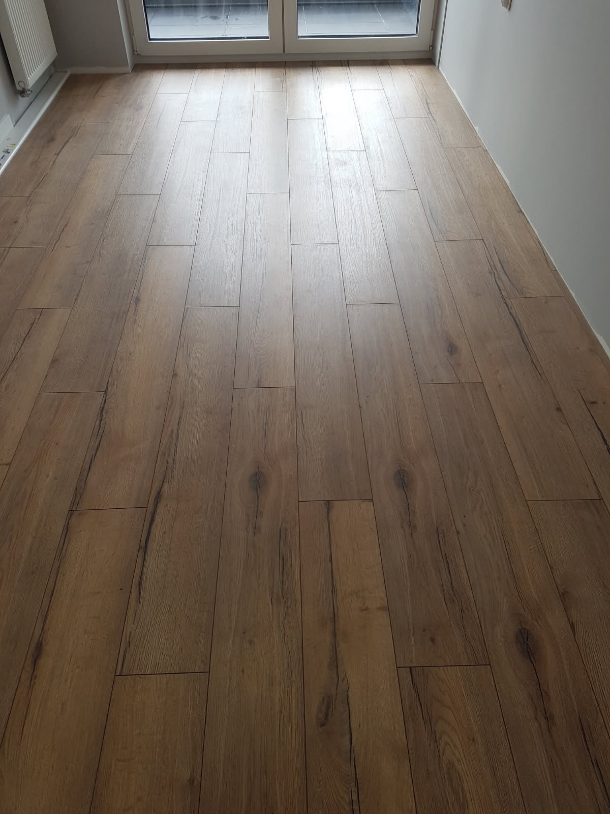 Ułożona podłoga z paneli imitujących drewno w jasnym odcieniu brązu, widok z góry, z oknem w tle. Minimalistyczne wykończenie wnętrza.