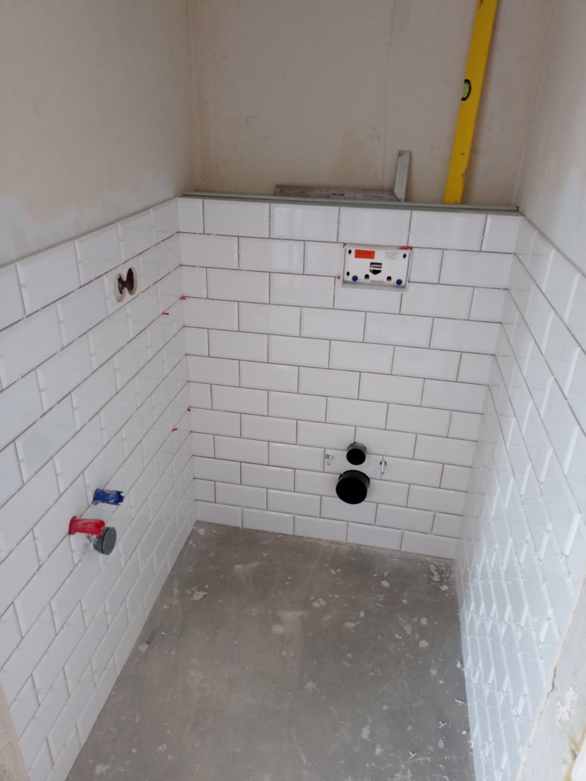 Ściany w trakcie wykańczania białymi, prostokątnymi płytkami. Widoczne rurki, poziomica i niezakończona podłoga. Małe krzyżyki dystansowe między płytkami.