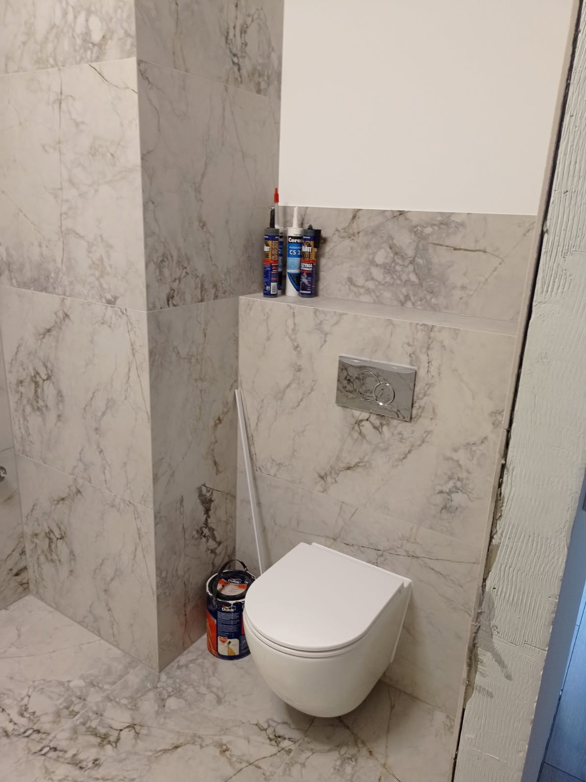 Nowoczesna łazienka z marmurowymi płytkami, wiszącą toaletą i srebrnym przyciskiem spłukującym. Widoczne tuby z silikonem i farba, sugerujące etap wykończeniowy.