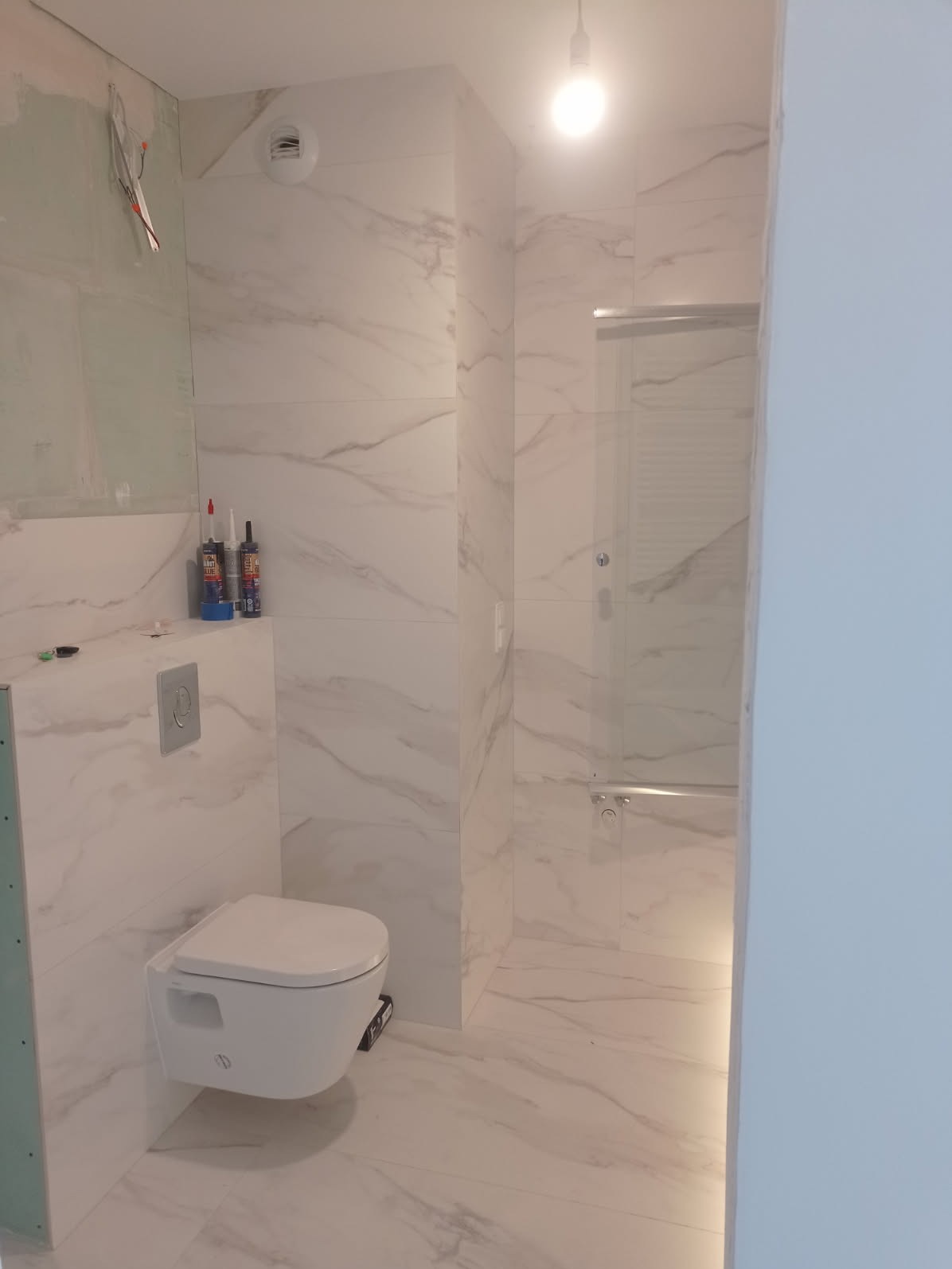 Wnętrze łazienki w trakcie wykańczania: białe płytki imitujące marmur, podwieszana toaleta, częściowo zamontowana kabina prysznicowa, widoczne materiały budowlane.