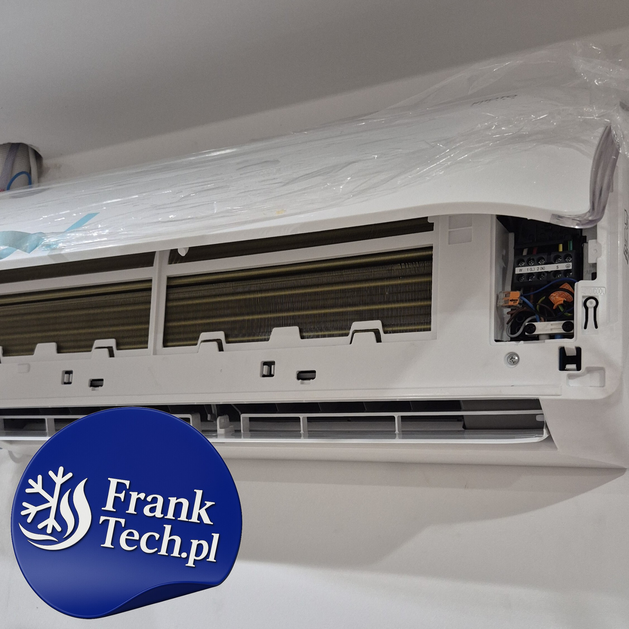 Biała klimatyzacja ścienna, jeszcze w folii ochronnej, z widocznymi elementami instalacji elektrycznej i logotypem firmy Frank Tech.pl w lewym dolnym rogu.