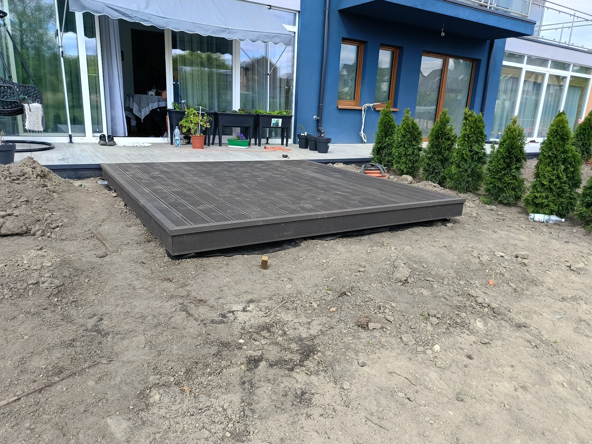 Montaż tarasu kompozytowego w Chorzowie. Widok na częściowo ułożony, brązowy taras z desek kompozytowych, z widocznym podłożem i roślinami w tle.