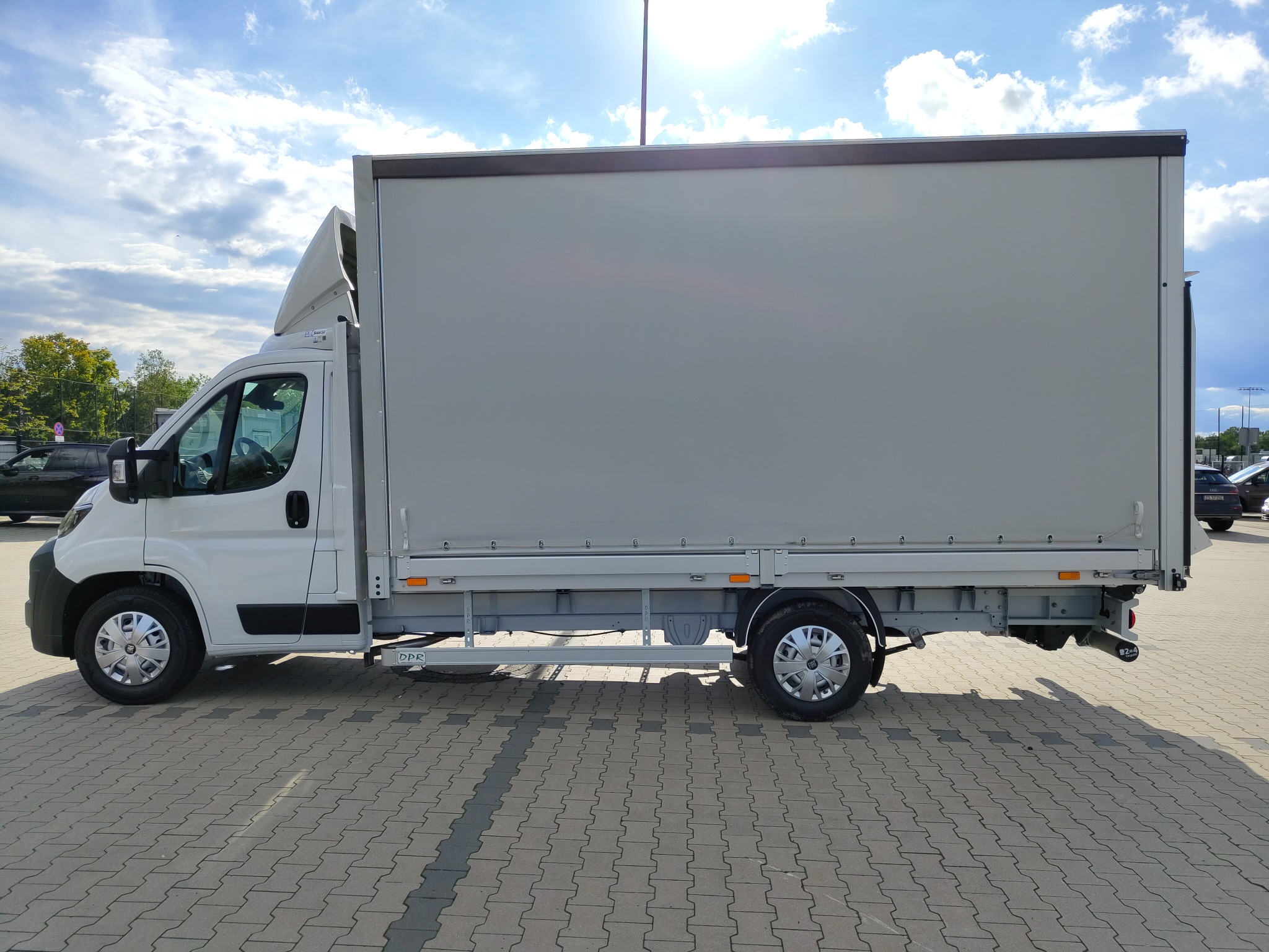 Biały samochód dostawczy z plandeką na parkingu z kostki, widok z boku, słoneczny dzień, błękitne niebo z chmurami.