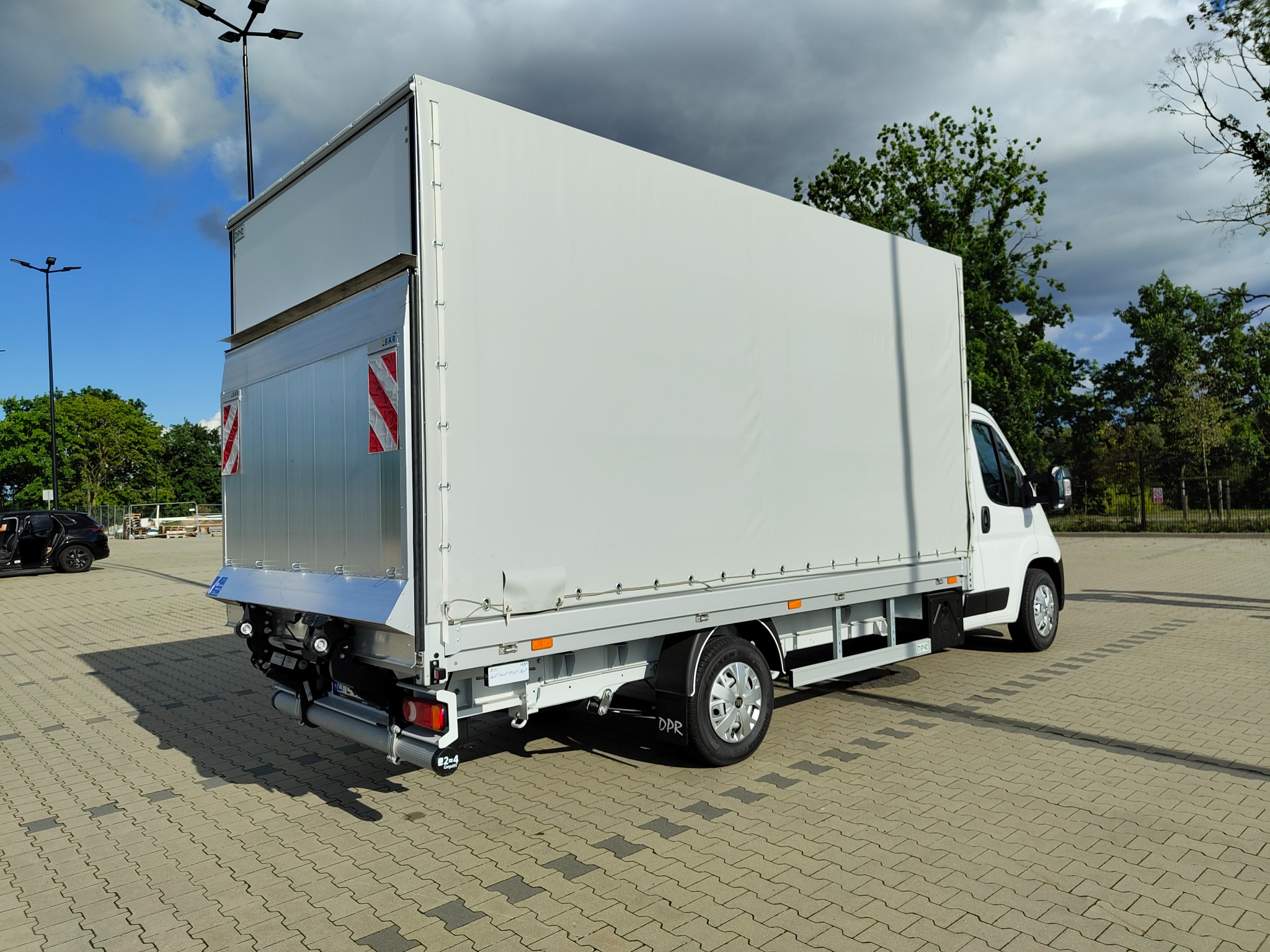 Biały samochód dostawczy z plandeką na parkingu, widok z tyłu. Czerwono-białe oznaczenia ostrzegawcze, opony z czarnymi chlapaczkami. Pochmurne niebo w tle.