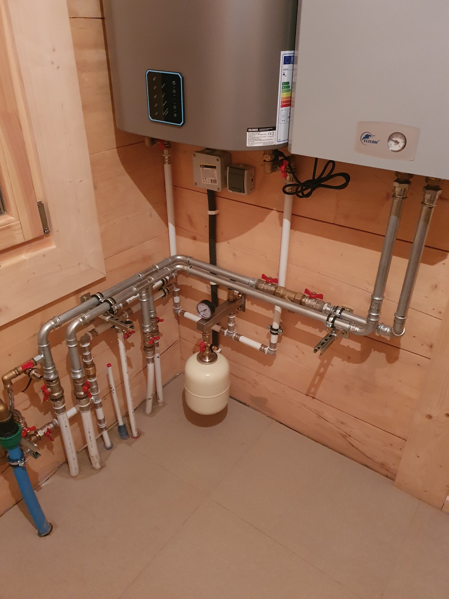 Złożona instalacja grzewcza z dwoma kotłami w drewnianym pomieszczeniu, widoczne liczne rury, zawory i zbiornik wyrównawczy. Całość zamontowana na drewnianej ścianie.