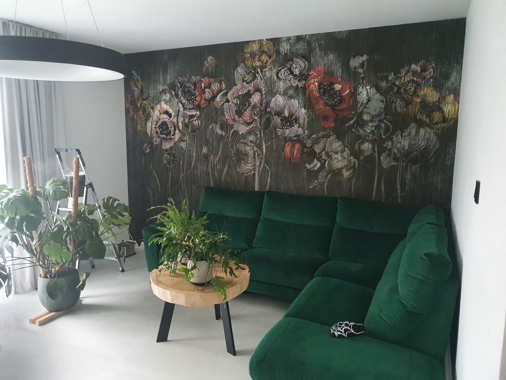 Pokój po wykończeniu: ściana z motywem kwiatowym, zielona sofa narożna, drewniany stolik kawowy z paprotką. W tle drabina malarska, lampa sufitowa.