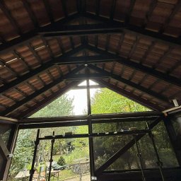 PAG OKNA I DRZWI Piotr Gniech - Wnętrze drewnianej altany z widokiem na zieleń przez trójkątne okno w dachu. Widoczne elementy konstrukcyjne i fragmenty wyposażenia.