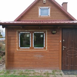 PAG OKNA I DRZWI Piotr Gniech - Drewniany domek z nowymi oknami PCV, zabezpieczonymi pianką montażową. Widoczna brązowa stolarka okienna i drzwiowa, czerwony dach i fragment trawnika.