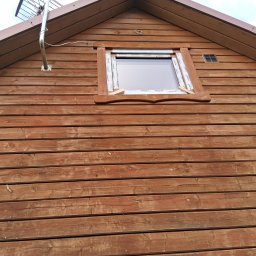 PAG OKNA I DRZWI Piotr Gniech - Drewniana elewacja domu z zamontowanym oknem. Widoczna antena na dachu i kratka wentylacyjna. Okno z ramą z tworzywa sztucznego i drewnianą obudową.