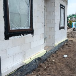 PAG OKNA I DRZWI Piotr Gniech - Surowy stan budynku z białych bloczków z zamontowanymi oknami z brązowymi ramami, zabezpieczonymi folią. Widoczna izolacja fundamentów i teren budowy.