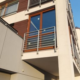 PAG OKNA I DRZWI Piotr Gniech - Nowoczesny balkon z drewnianą ramą okienną i metalową balustradą, widok z dołu na tle błękitnego nieba. Elementy architektoniczne budynku w jasnych kolorach.
