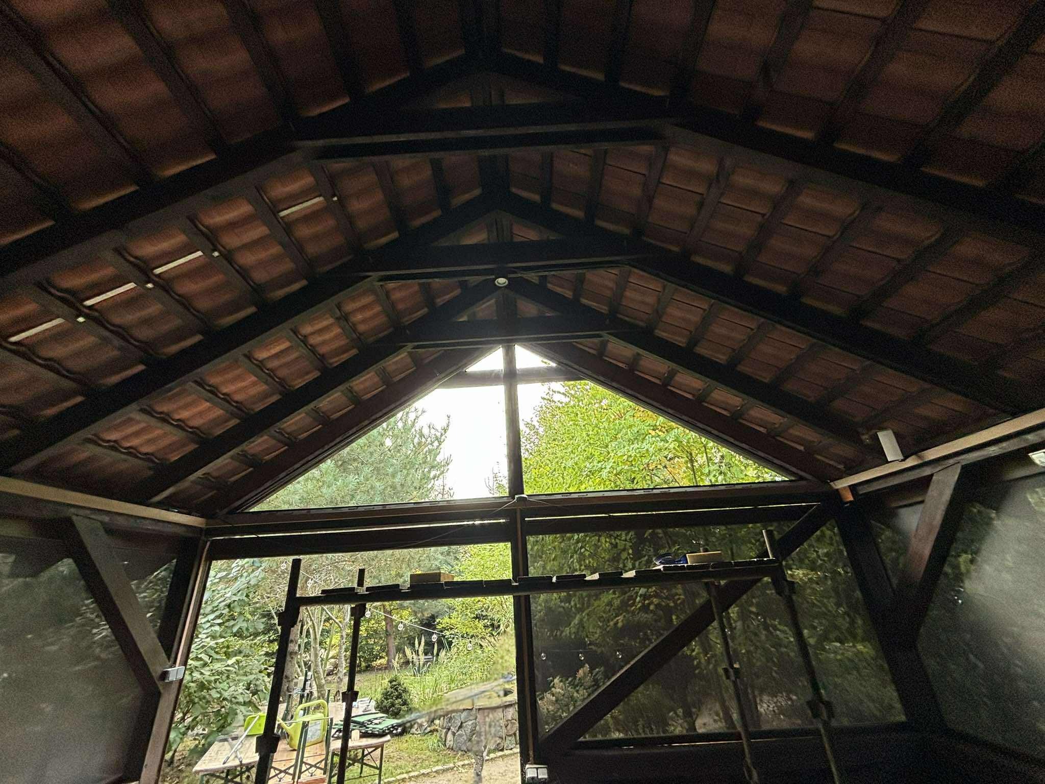 Wnętrze drewnianej altany z widokiem na zieleń przez trójkątne okno w dachu. Widoczne elementy konstrukcyjne i fragmenty wyposażenia.