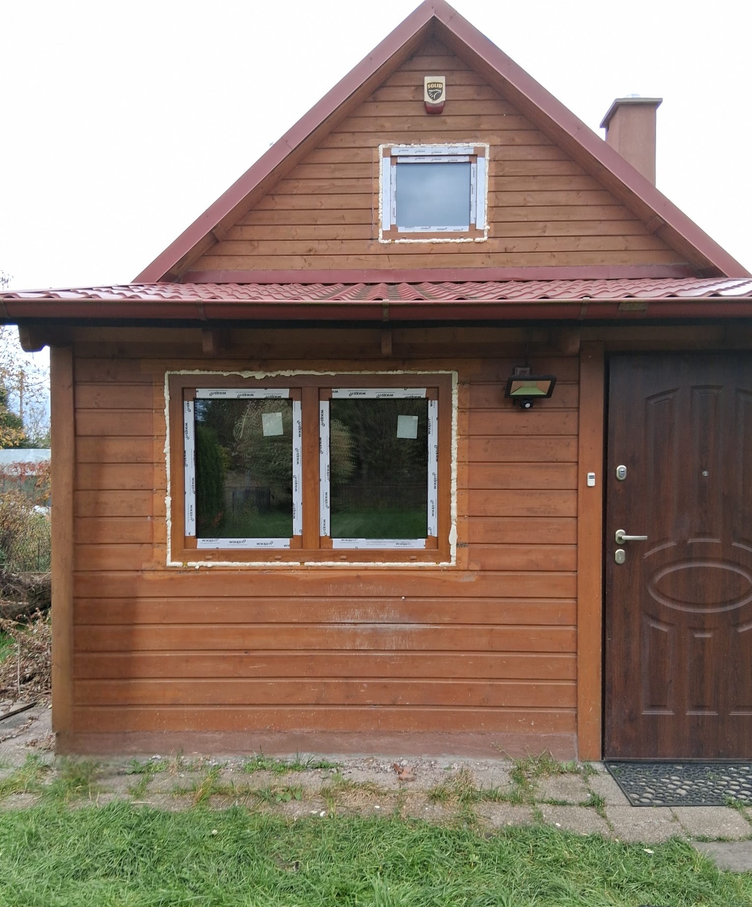Drewniany domek z nowymi oknami PCV, zabezpieczonymi pianką montażową. Widoczna brązowa stolarka okienna i drzwiowa, czerwony dach i fragment trawnika.