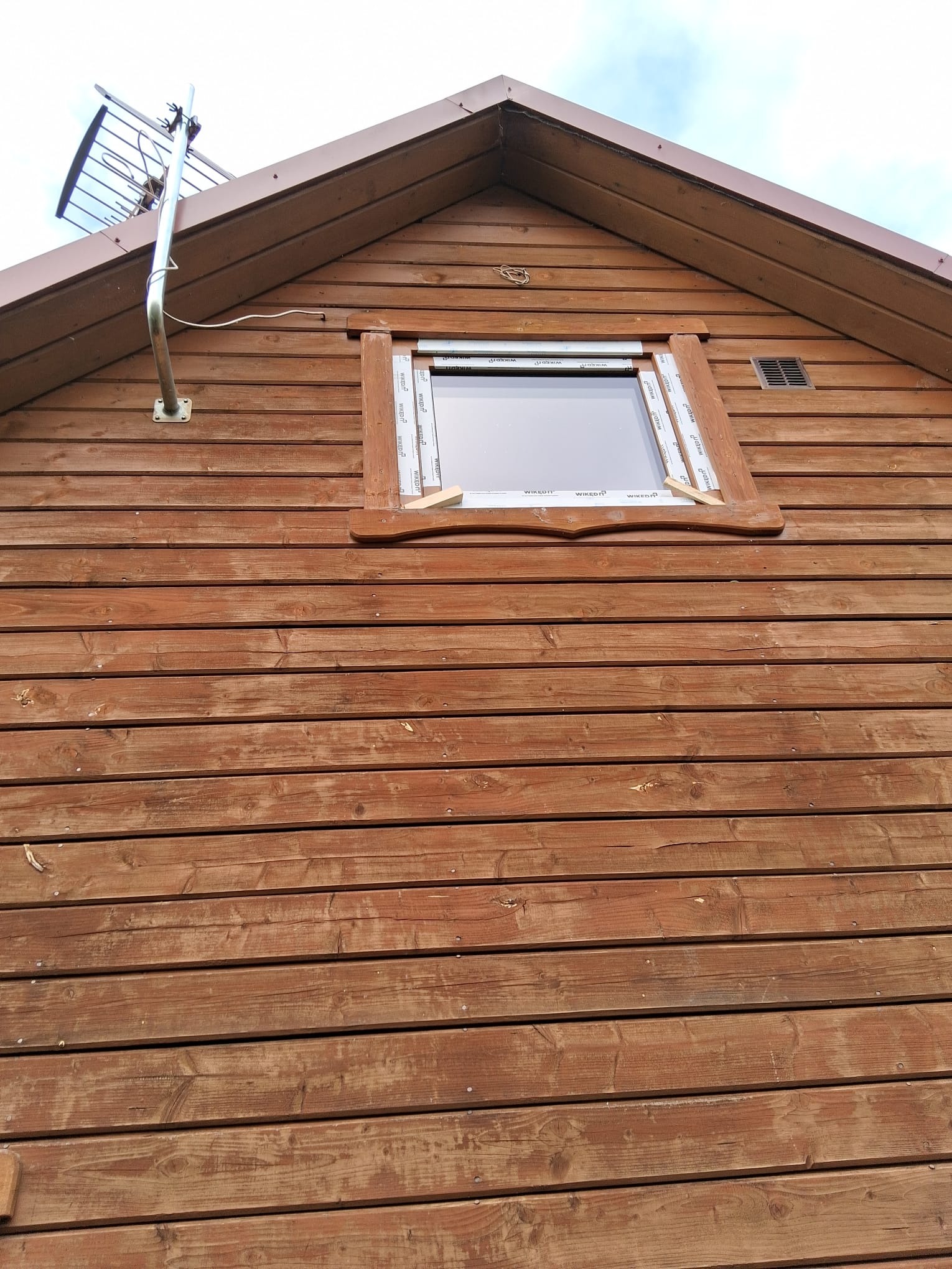 Drewniana elewacja domu z zamontowanym oknem. Widoczna antena na dachu i kratka wentylacyjna. Okno z ramą z tworzywa sztucznego i drewnianą obudową.