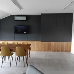 Nowoczesna aranżacja wnętrza z zabudową pod skosem: antracytowe szafy na wymiar, drewniana komoda, stół z krzesłami. Widoczny klimatyzator i drabina malarska.