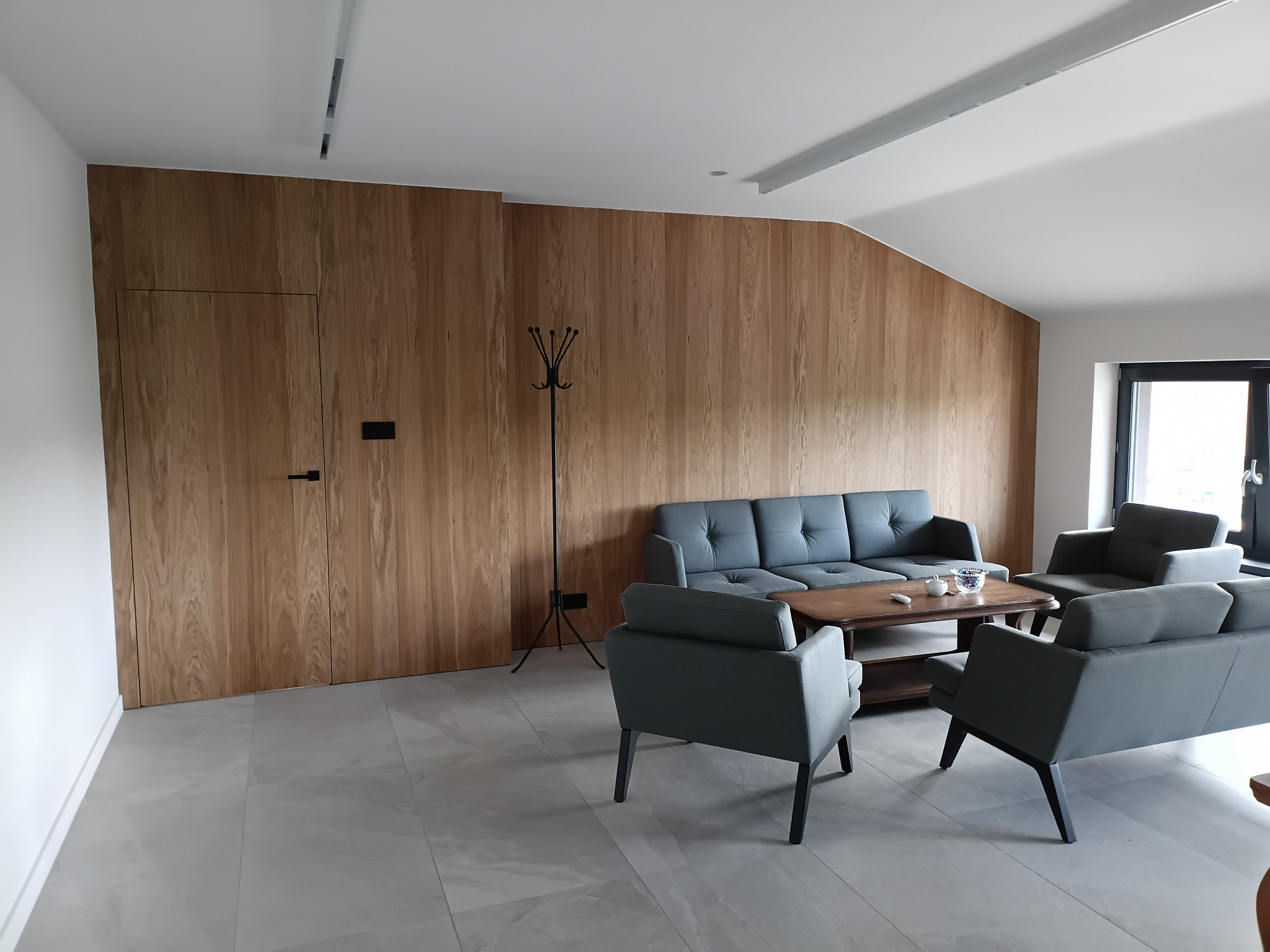 Nowoczesny salon z ścianą wyłożoną drewnianymi panelami, ukrywającymi drzwi. Szara sofa i fotele ustawione wokół drewnianego stolika kawowego. Minimalistyczny design.