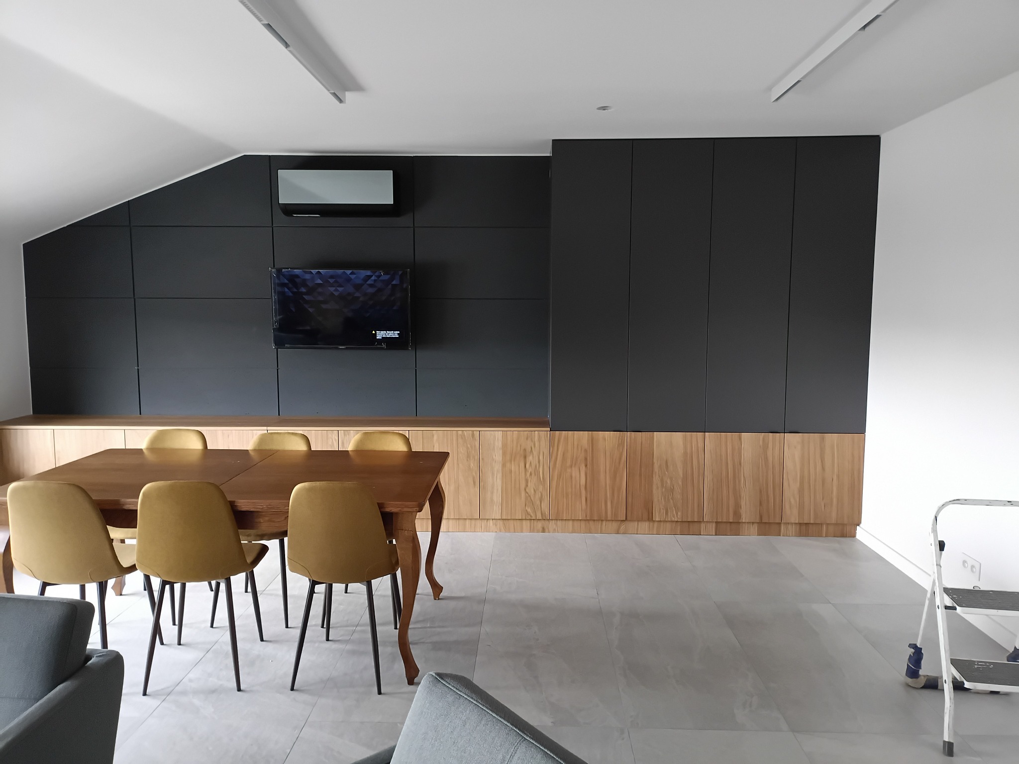 Nowoczesna aranżacja wnętrza z zabudową pod skosem: antracytowe szafy na wymiar, drewniana komoda, stół z krzesłami. Widoczny klimatyzator i drabina malarska.