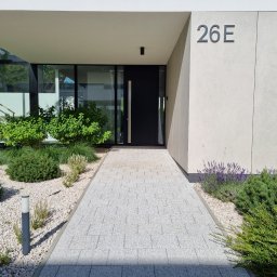 Zielony Styl - Nowoczesne wejście do domu z jasną kostką brukową, rabatami z kamyków i zielenią. Minimalistyczny design z numerem domu '26E' i czarnymi drzwiami.