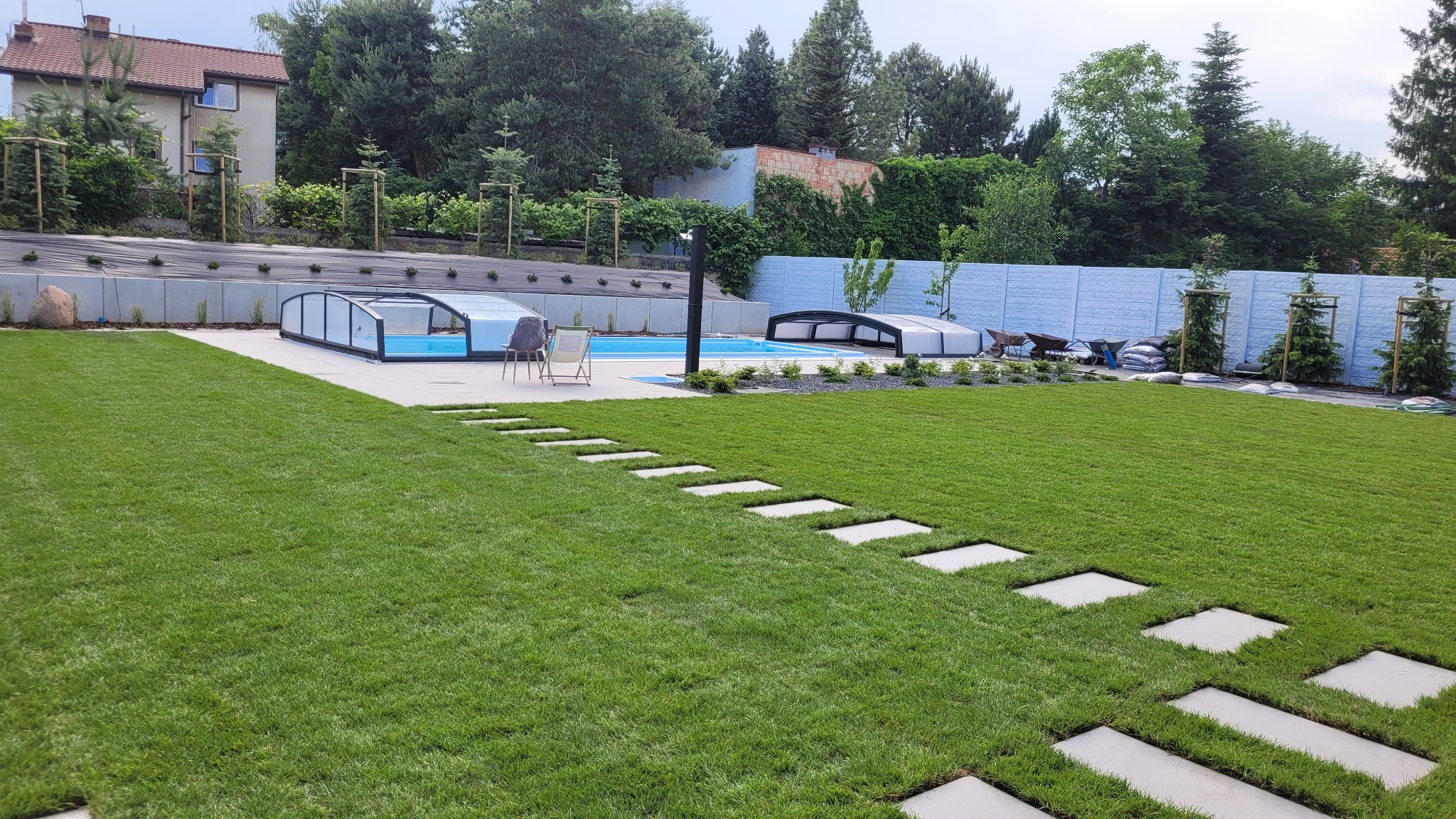 Nowoczesny ogród z basenem i ścieżką z płyt na trawniku. W tle jasny płot i drzewa. Widoczne leżaki i zadaszenia basenowe. Estetyczne zagospodarowanie przestrzeni.