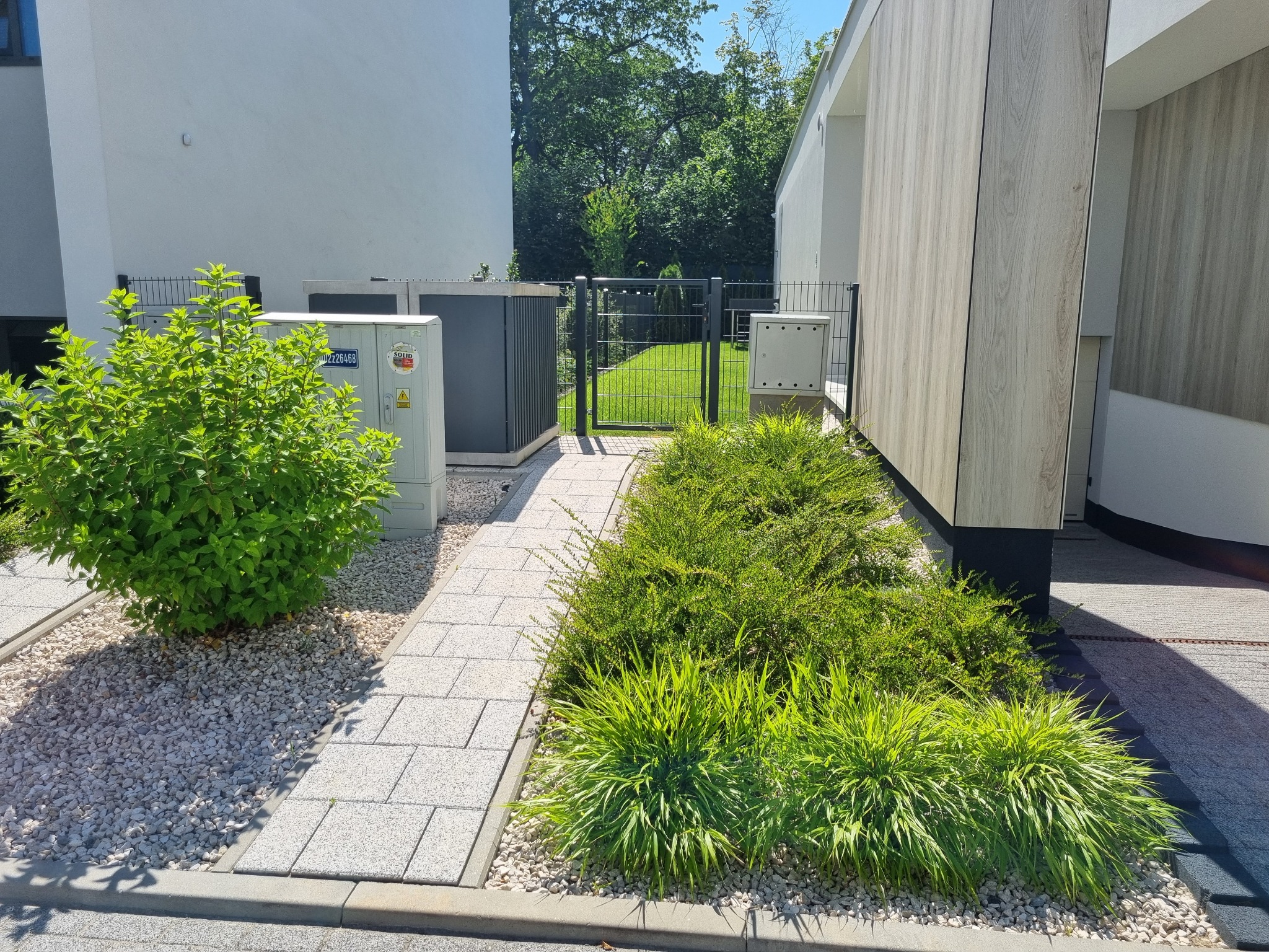 Nowoczesny ogród z kostką brukową, ozdobnym żwirem i zielenią, w otoczeniu minimalistycznej architektury budynków. Widok na furtkę i trawnik.