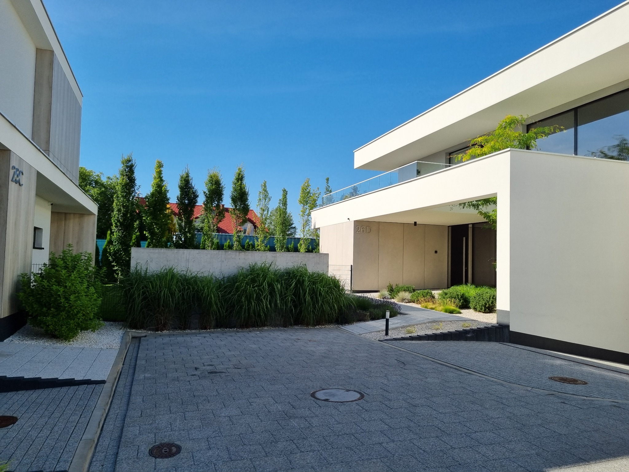 Nowoczesny dom z brukowanym podjazdem i minimalistycznym ogrodem. Widoczne geometryczne kształty budynków i starannie dobrane rośliny. Jasne barwy i błękitne niebo.