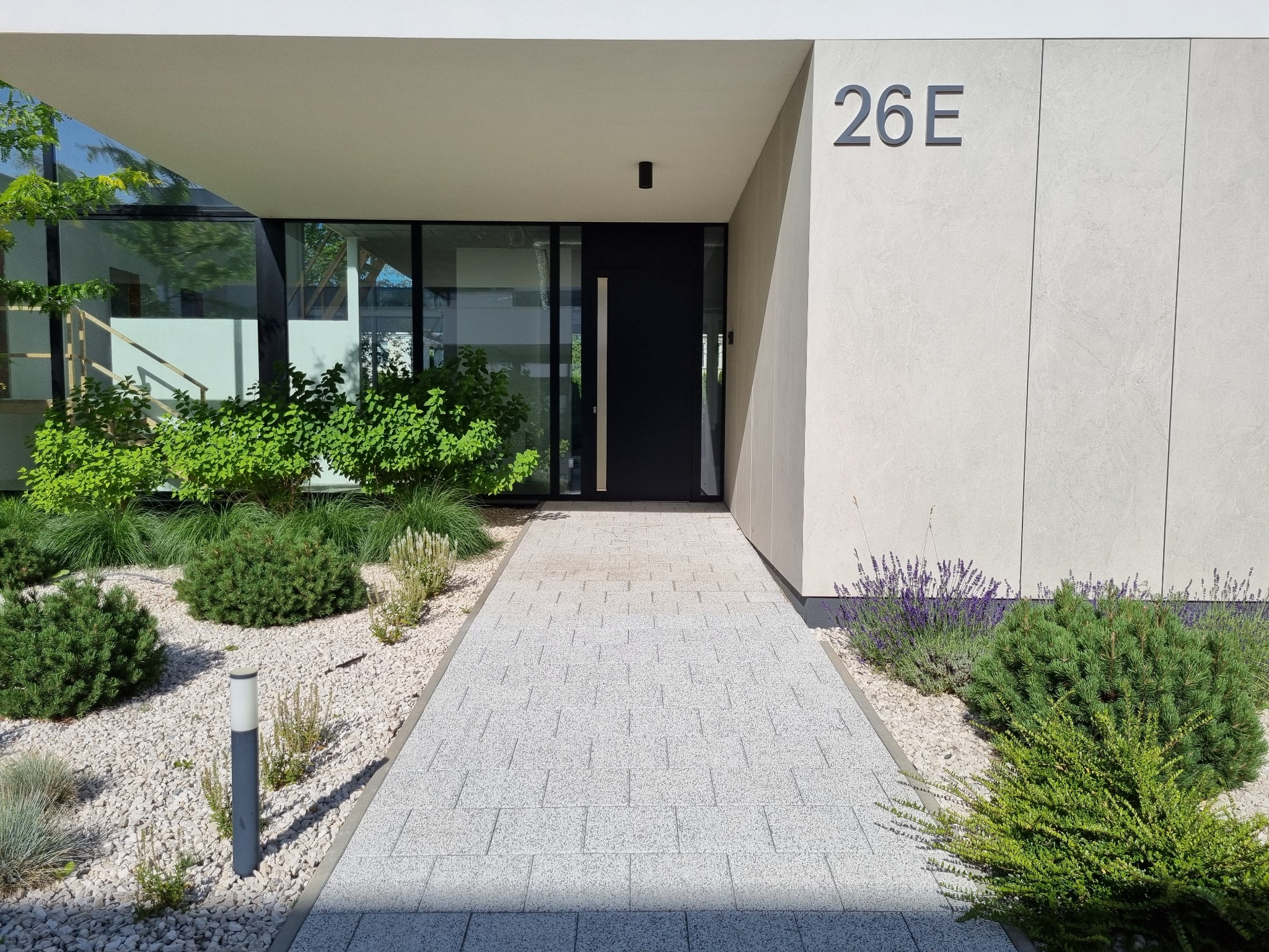 Nowoczesne wejście do domu z jasną kostką brukową, rabatami z kamyków i zielenią. Minimalistyczny design z numerem domu '26E' i czarnymi drzwiami.