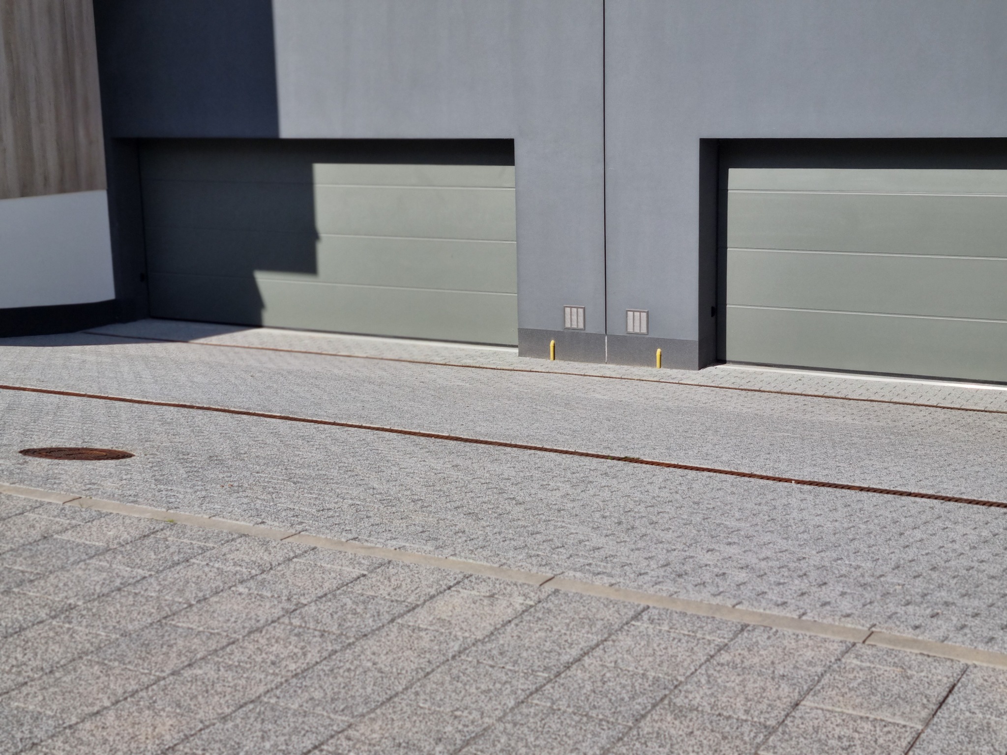 Szara kostka brukowa na podjeździe do garażu z dwiema bramami. Widoczne linie dylatacyjne i studzienka kanalizacyjna. Nowoczesny styl.