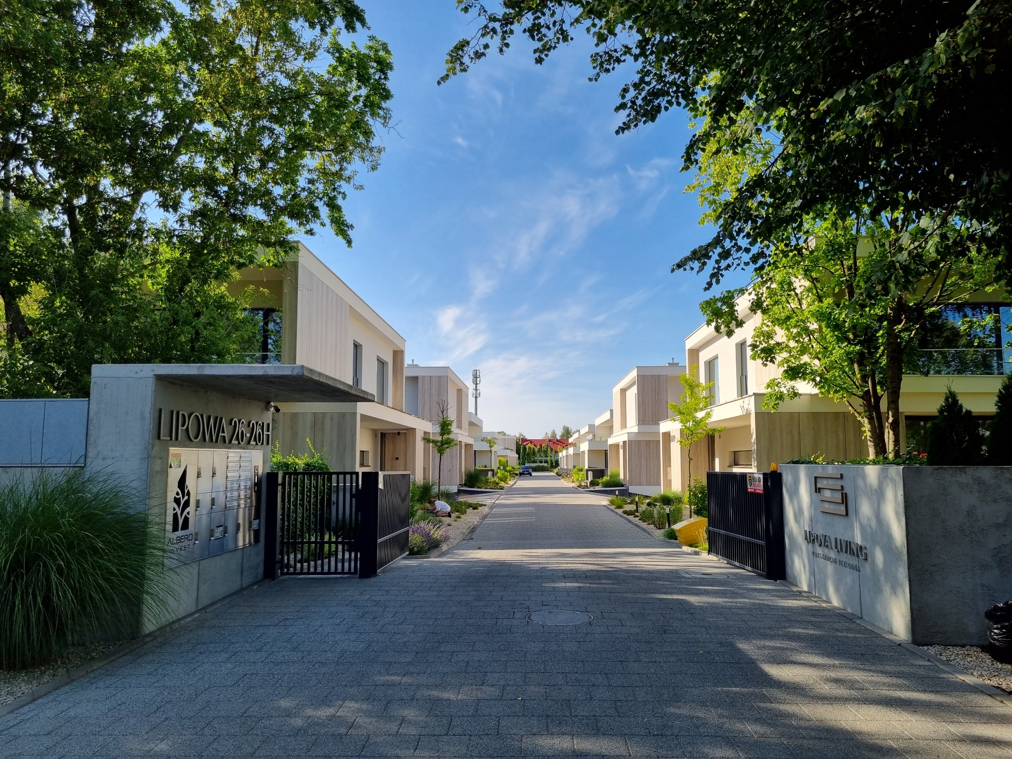 Osiedle Lipowa Living: aleja z kostki brukowej, nowoczesne domy z jasną elewacją, zadbana zieleń i błękitne niebo.