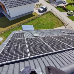 Grabka.IT - Tomasz Grabka - Panele słoneczne na szarym dachu domu jednorodzinnego, widok z góry. W tle trawnik, ogrodzenie i zaparkowane samochody. Część kadru zajmują stopy osoby montującej.