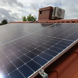 Grabka.IT - Tomasz Grabka - Panele fotowoltaiczne na dachu z dachówki ceramicznej, obok komin z klimatyzatorem. Widok z dołu na pochmurne niebo i drzewa w tle. Instalacja paneli.