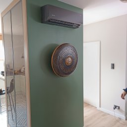 Grabka.IT - Tomasz Grabka - Szary klimatyzator Rotenso na zielonej ścianie z dekoracyjnym, okrągłym elementem. Widoczny fragment przeszklonej szafy i jasna podłoga.