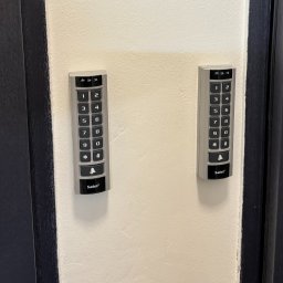 Grabka.IT - Tomasz Grabka - Dwa panele kodowe Satel zamontowane na ścianie obok ciemnych drzwi. Panele mają klawiaturę numeryczną i przyciski funkcyjne.
