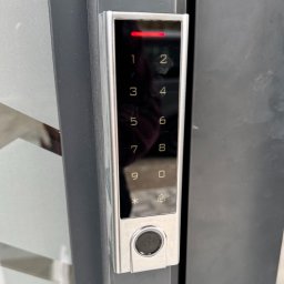 Grabka.IT - Tomasz Grabka - Nowoczesny panel dotykowy alarmu domowego z podświetlanymi cyframi i czytnikiem linii papilarnych, zamontowany na ciemnej powierzchni. Czerwona dioda sygnalizacyjna u góry.