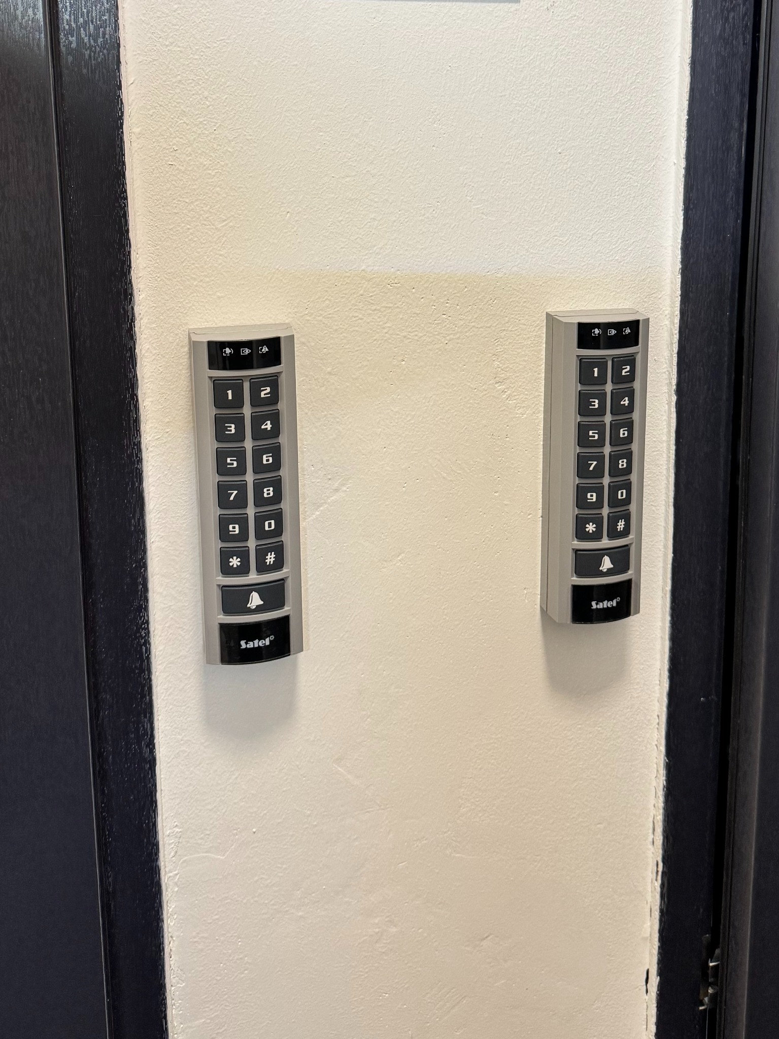 Dwa panele kodowe Satel zamontowane na ścianie obok ciemnych drzwi. Panele mają klawiaturę numeryczną i przyciski funkcyjne.