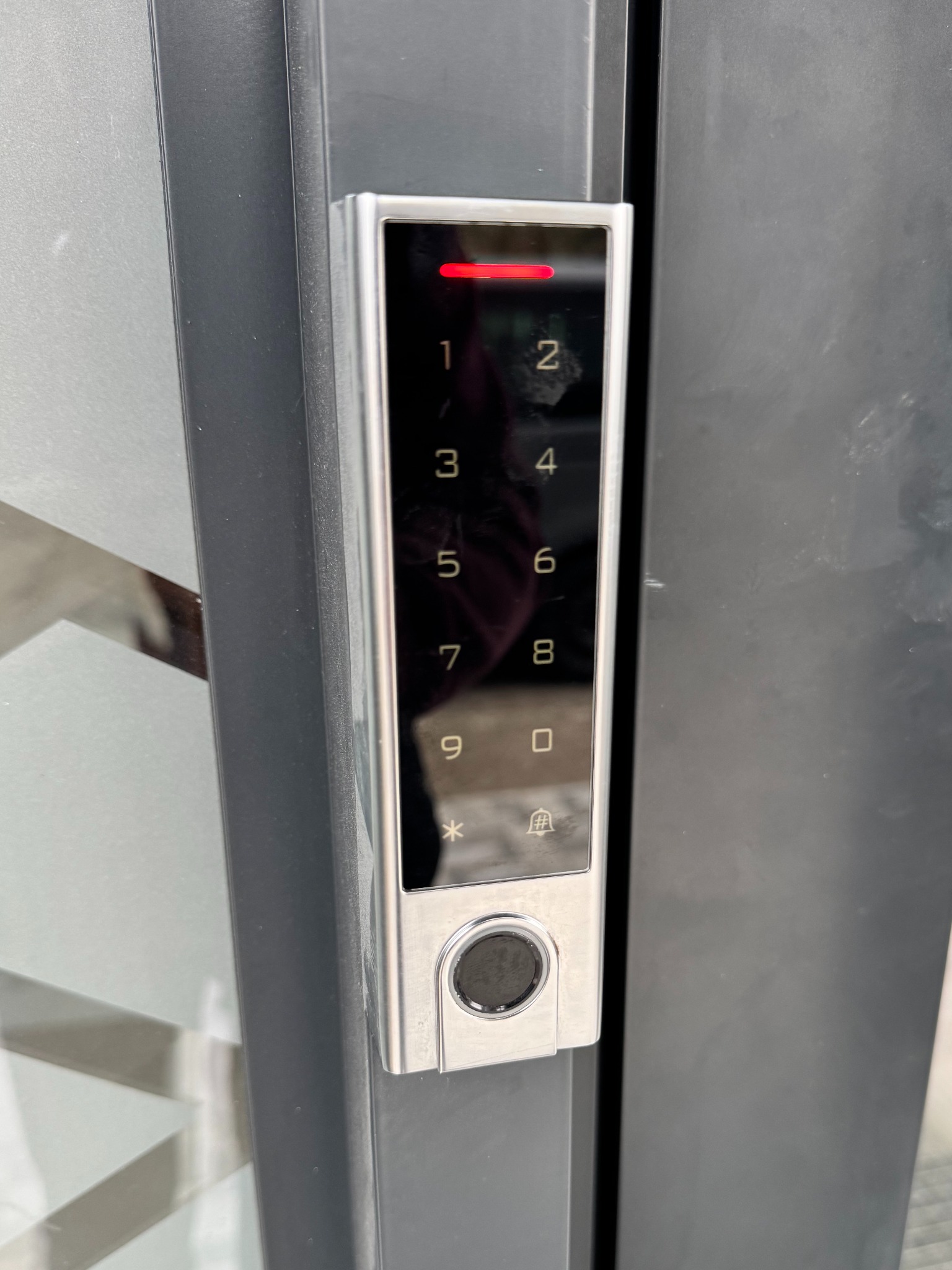 Nowoczesny panel dotykowy alarmu domowego z podświetlanymi cyframi i czytnikiem linii papilarnych, zamontowany na ciemnej powierzchni. Czerwona dioda sygnalizacyjna u góry.