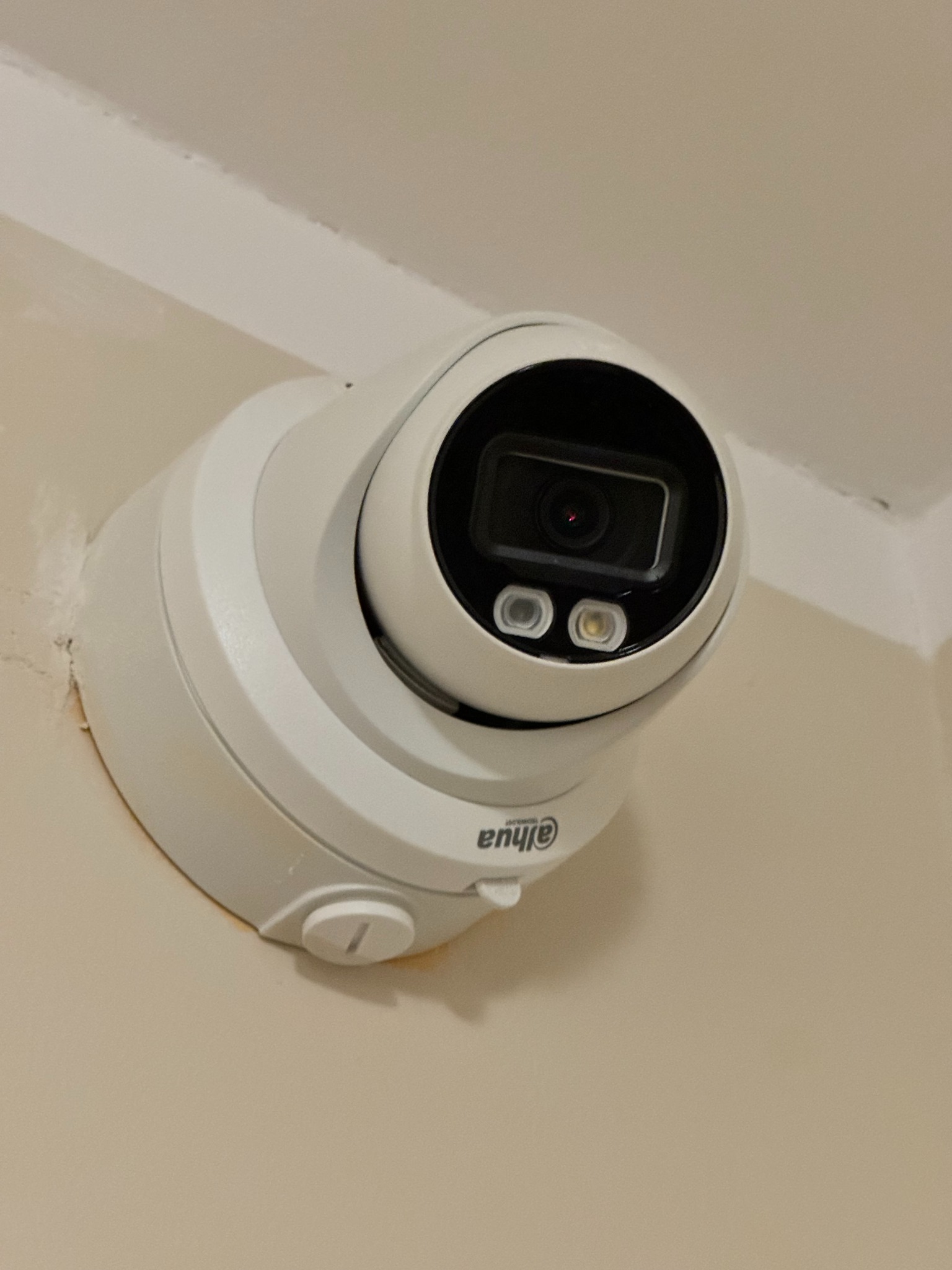 Zamontowana biała kamera kopułkowa Dahua z widocznymi diodami IR, przymocowana do ściany w rogu pomieszczenia. Widok z dołu, bliska perspektywa.