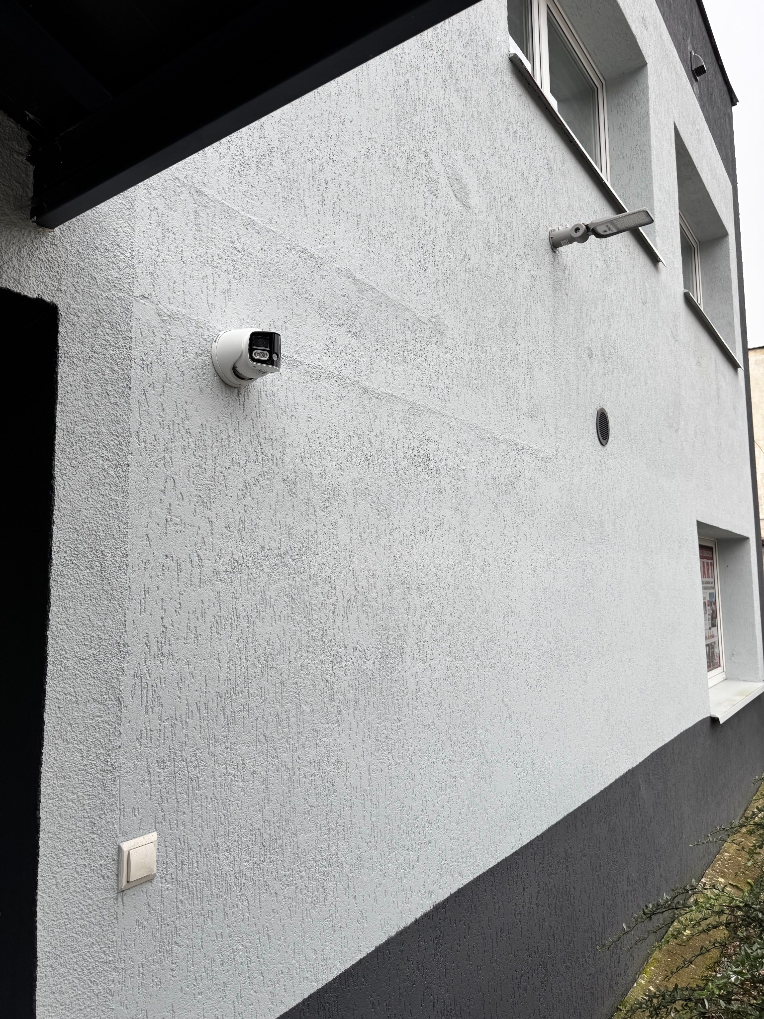 Biała kamera monitoringu na elewacji budynku z szarym cokołem i oknami. Widoczny także włącznik światła i lampa zewnętrzna. Ujęcie z dołu.