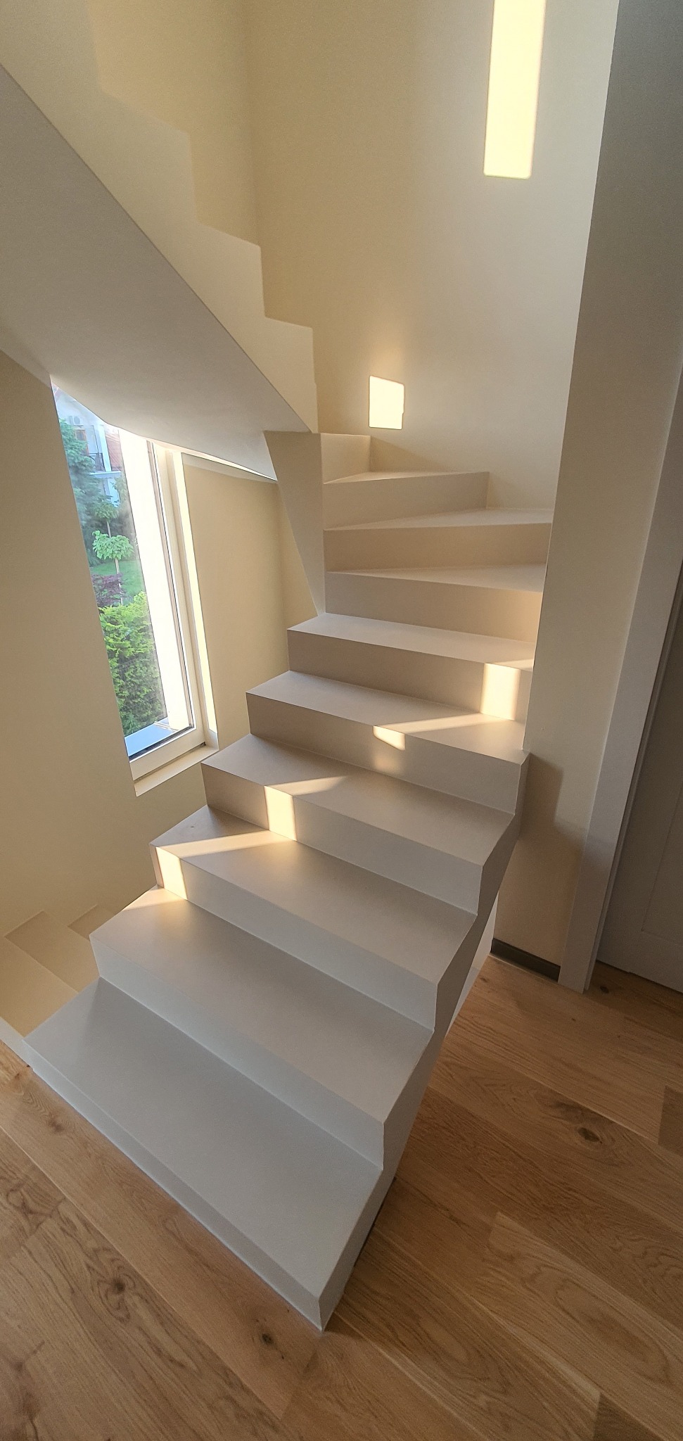 Nowoczesne, białe schody z subtelnym oświetleniem, prowadzące do góry. Drewniana podłoga i okno z widokiem na zieleń. Minimalistyczny design, ciepłe światło.