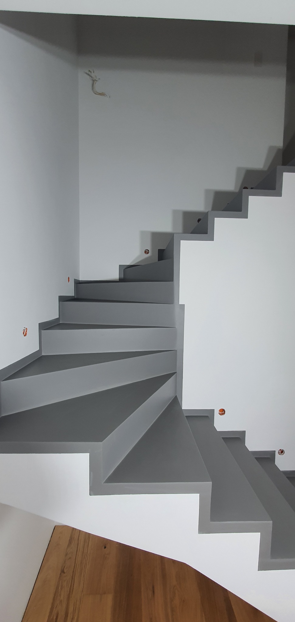 Nowoczesne, spiralne schody w odcieniach szarości i bieli, z widocznymi puszkami elektrycznymi na ścianie i fragmentem drewnianej podłogi.