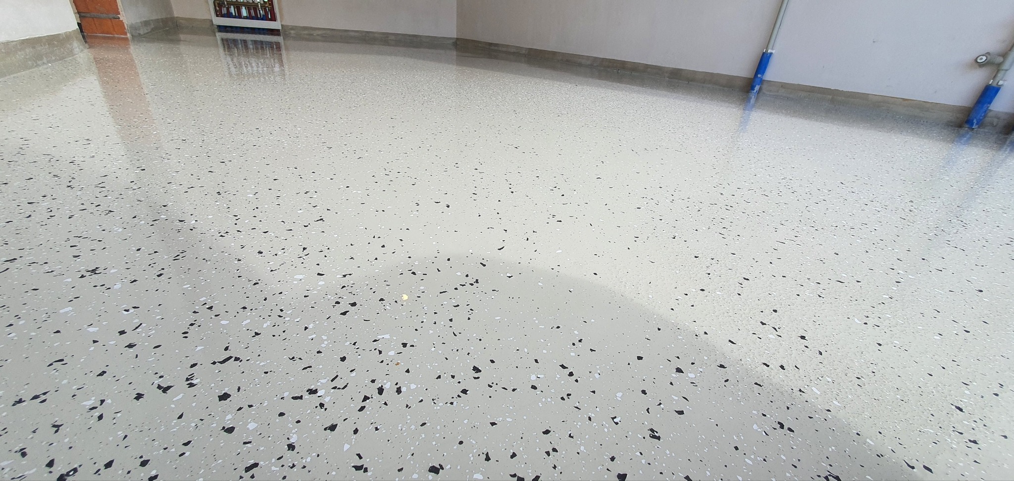 Błyszcząca, jasna posadzka z drobnymi, ciemnymi i białymi elementami dekoracyjnymi, odbijająca światło w garażu. Widoczne rury i drzwi w tle.