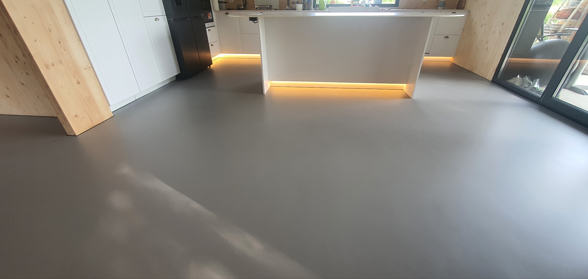 Szara, gładka posadzka w nowoczesnej kuchni z wyspą, oświetleniem LED pod szafkami i dużym oknem. Minimalistyczny design, czyste linie, jasne barwy.