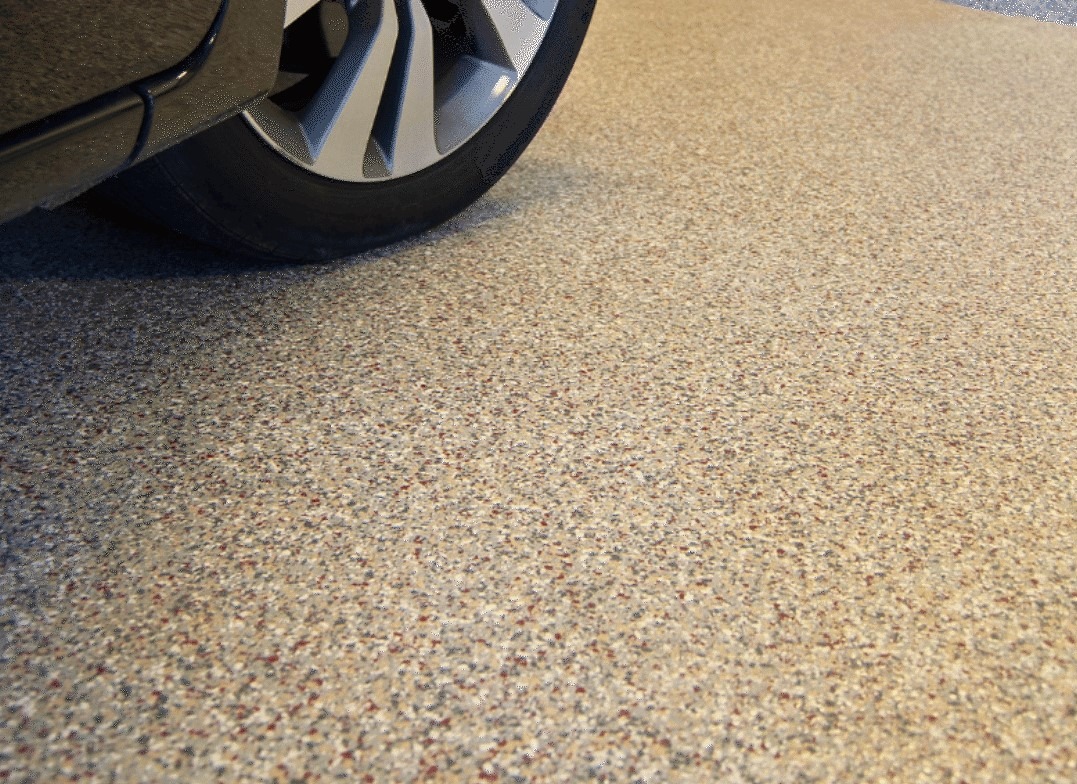 Beżowa posadzka żywiczna z drobnymi kamyczkami w garażu, z fragmentem koła samochodu w lewym górnym rogu.