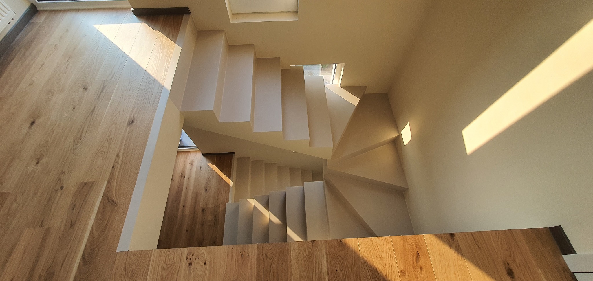 Minimalistyczne schody z mikrocementu, widok z góry, naturalne światło podkreśla fakturę. Drewniana podłoga i stopnie tworzą harmonijną całość.