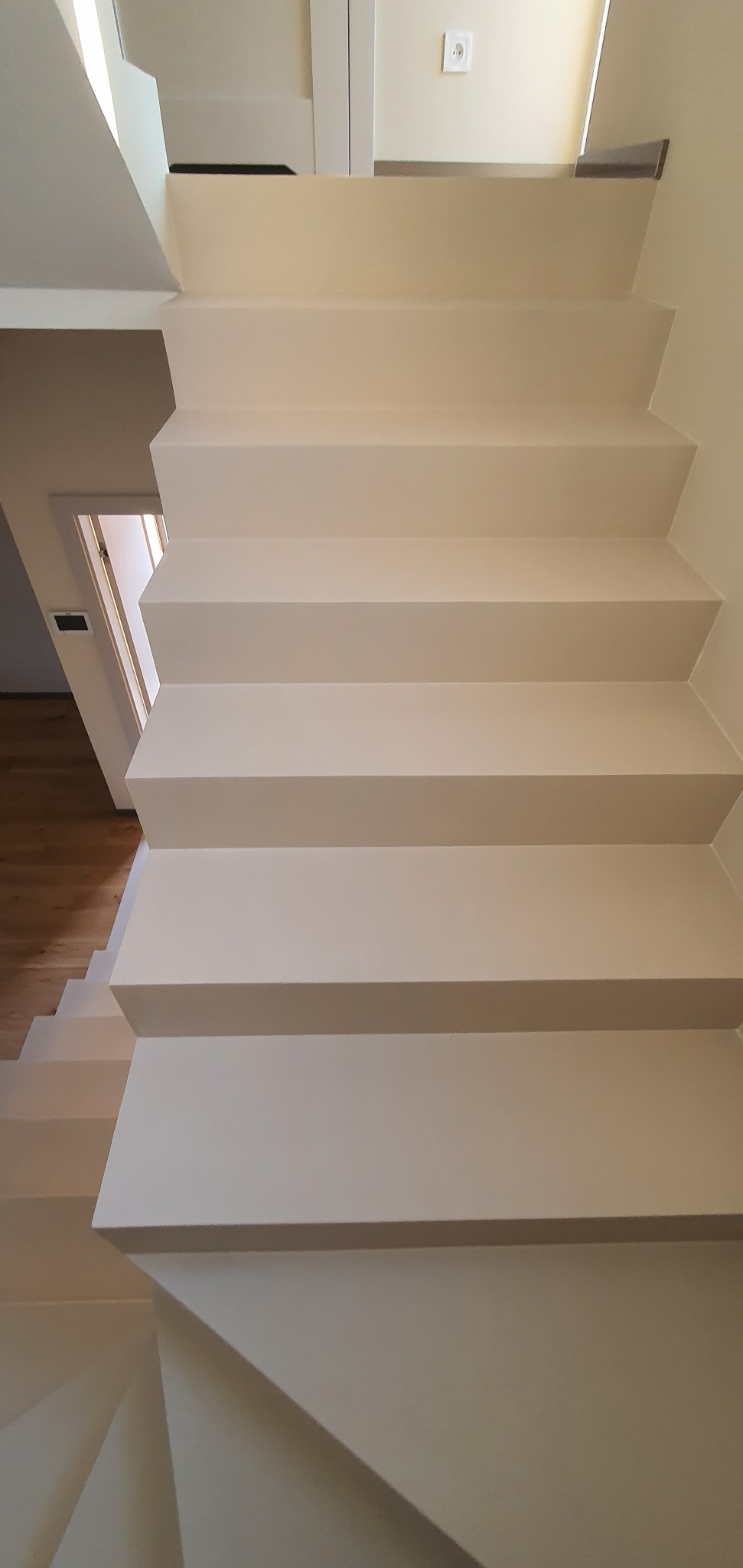 Kremowe schody wewnętrzne z równymi stopniami, widoczne z dołu, z otwartymi drzwiami i drewnianą podłogą w tle. Minimalistyczny design.