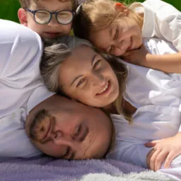 Uśmiechnięta rodzina w białych koszulach leżąca na kocu na trawie, z bliska, w słoneczny dzień. Dzieci przytulają się do rodziców. Wesoła atmosfera.