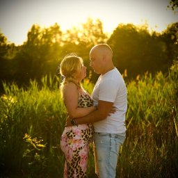 Passion.photos - Zakochana para, kobieta w kwiecistej sukience ciążowej, mężczyzna w białej koszulce, obejmują się na tle zieleni i zachodzącego słońca. Romantyczna sesja w plenerze.