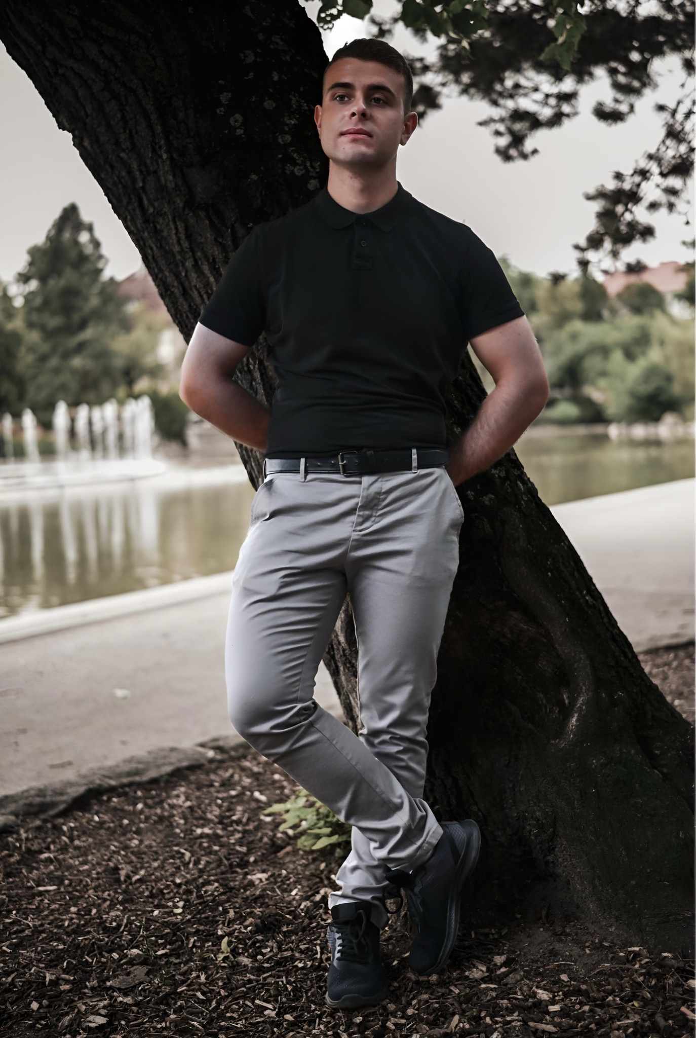 Młody mężczyzna w czarnej koszulce polo i szarych spodniach, oparty o drzewo w parku z fontanną w tle, pozujący do zdjęcia.