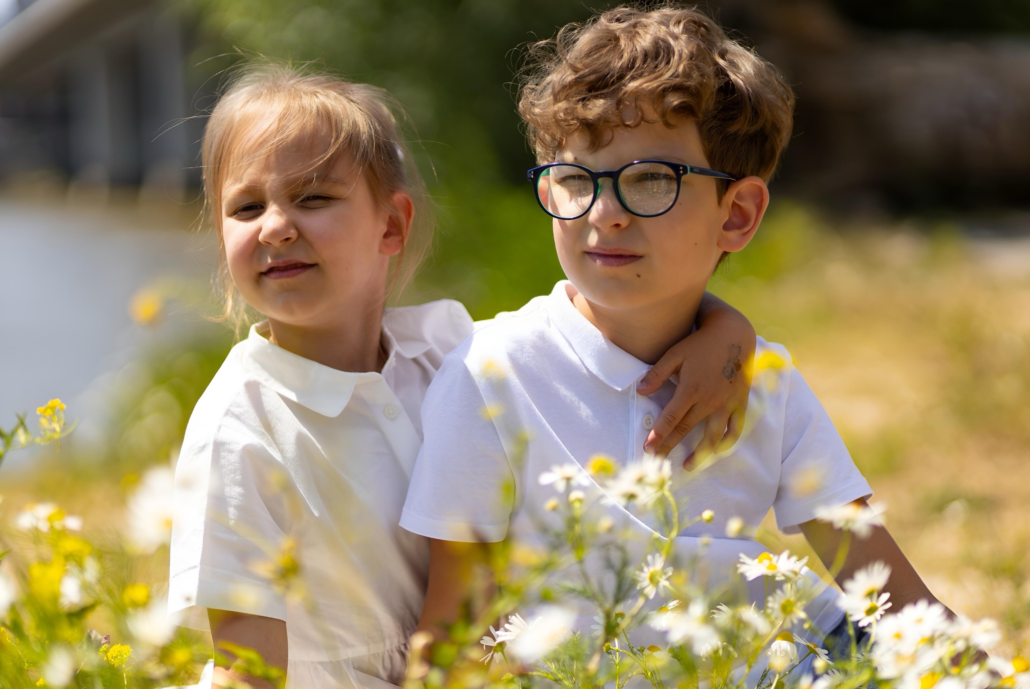 Portret dwójki dzieci w białych ubraniach, chłopiec w okularach obejmuje dziewczynkę na tle kwitnącej łąki z żółtymi i białymi kwiatami w słoneczny dzień.