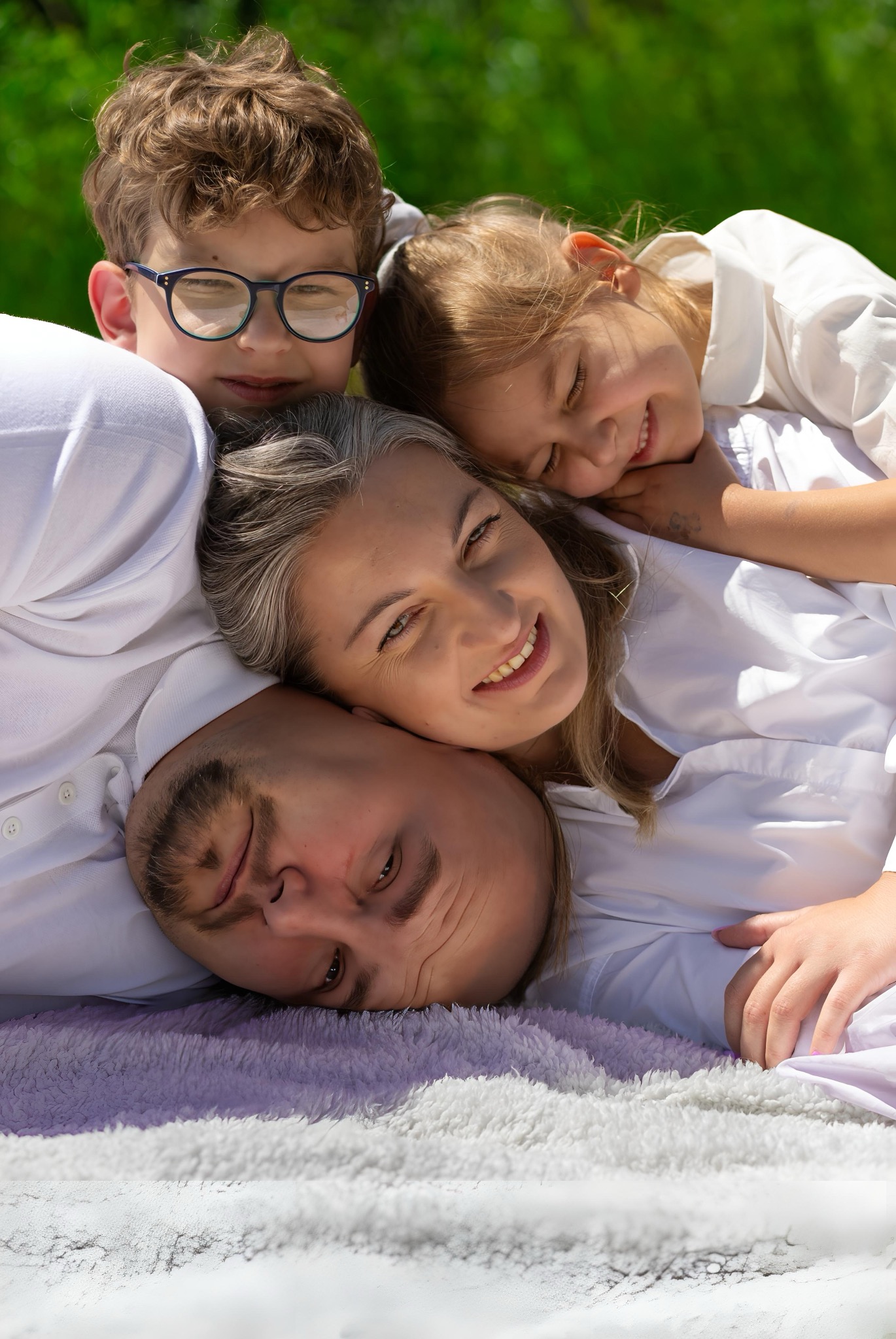 Uśmiechnięta rodzina w białych koszulach leżąca na kocu na trawie, z bliska, w słoneczny dzień. Dzieci przytulają się do rodziców. Wesoła atmosfera.