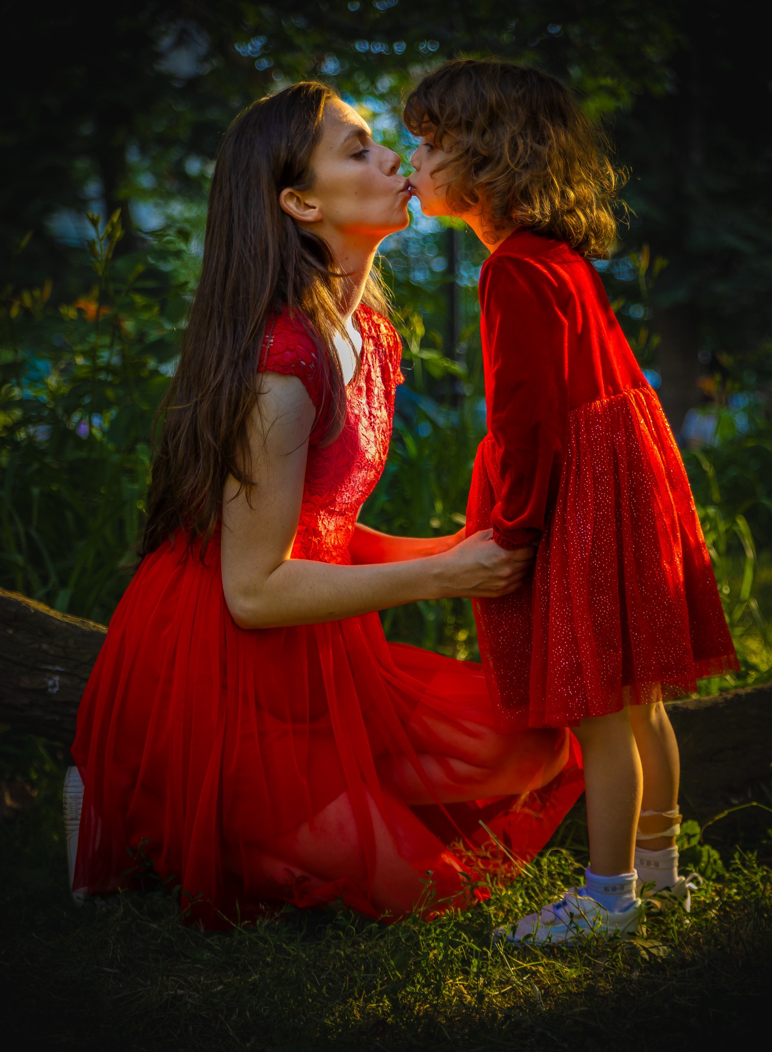 Matka w czerwonej sukni całuje córkę, również w czerwonej sukience, w plenerze, na tle zieleni. Ujęcie w kontraście, z ciemnym tłem i jasnymi postaciami.